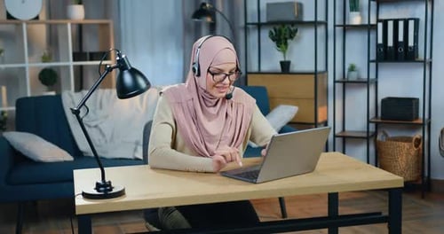 Mujer árabe en hiyab sentada frente a una computadora portátil con auriculares durante una videoconferencia en línea