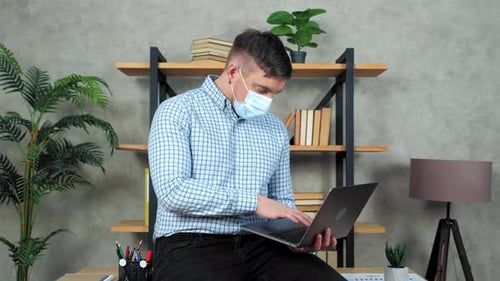 Businessman in protective medical mask on face sits at desk in office use laptop