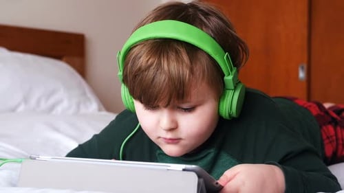 Child Wearing Headphones Uses Tablet on Bed