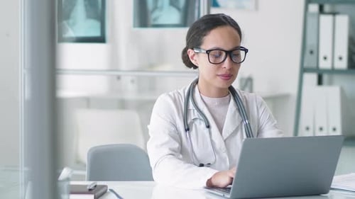 Female Doctor Using Laptop and Smiling at Camera at Workplace in Clinic