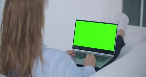 A Young Woman Lying on the Couch Looks at the Screen of the Laptop with a Green Screen and Nods Her