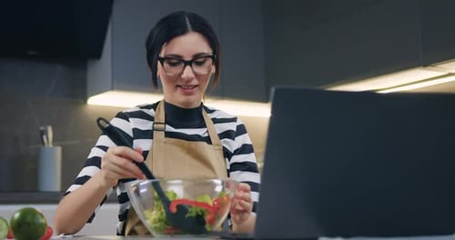 Mujer con gafas viendo una película en el ordenador portátil y mezclando ensalada de verduras en un tazón con una cuchara