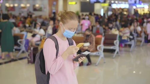 A Young Woman Wearing a Medical Face Mask in an Airport Stands in a Hall with a Cellphone in Her