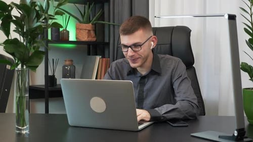 Man in earphones is doing online video chat conference sitting at office desk and looking at laptop