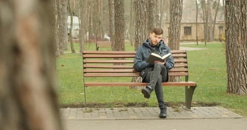Junger bärtiger Mann liest ein Buch auf einer Bank im Herbstpark in blauer Jacke