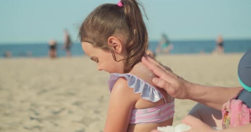 Mutter trägt Sonnenschutzlotion auf ihre kleine Tochter Haut auf, während sie am Strand sitzt