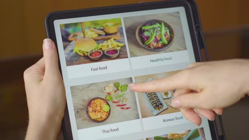 Young Woman Orders Food for Lunch Online Using a Tablet. She Is Sitting in a Kitchen in Her