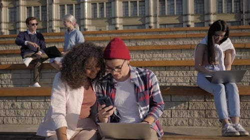 Beautiful Couple Uses the Phone on a Bench Near College. Students After Classes.