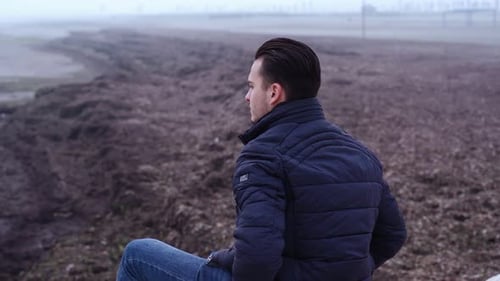 Pensive Man Sits on Bare Beach Stones at Vast Grey Sea