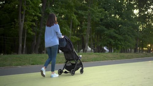 A Young Single Mother Walks with a Baby in a Stroller in the Park