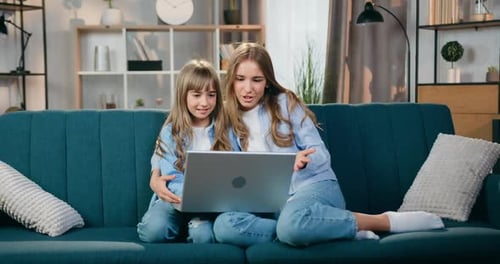 Amables dos hermanas rubias sentadas en un sofá suave en casa durante una videollamada positiva en una computadora portátil