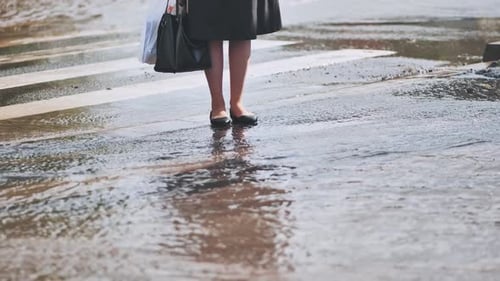 A Woman Stands on the Road and Tries to Cross the Road and Step Over Puddles After a Heavy Rain