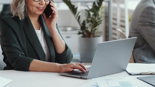 Mature Businesswoman Speaking on Phone and Typing on Laptop in Office