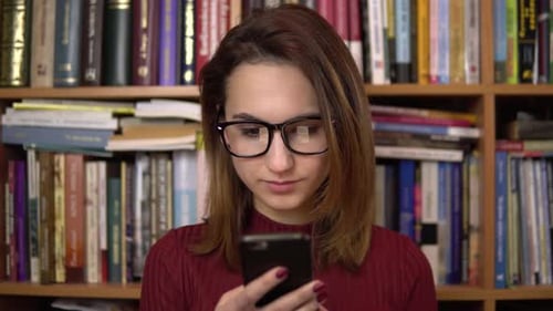 A Young Woman Is Looking at a Smartphone in a Library. Woman with Glasses Carefully Looks at the