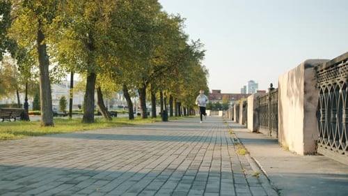 Active Young Man Jogging in Modern City and Listening to Music Through Headphones in Summer