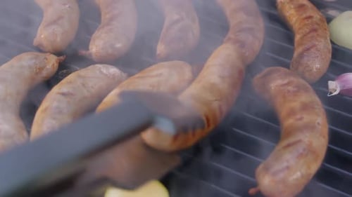 Mit Hilfe einer Metallzange werden die kochenden Würste auf einem schwarzen Barbecue-Grill von Hand gedreht