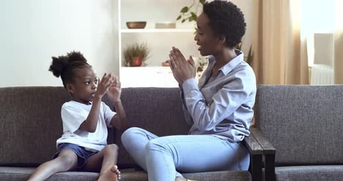 Mom and Daughter of African Ethnicity Clap Their Hands Sitting on Sofa at Home in Living Room