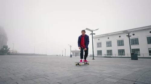 Young Man in Purple Jacket Skates or Rides Longboard on City Square in an Autumn Foggy Day Day
