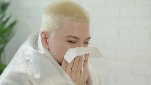 Une femme bouleversée aux cheveux blancs courts enveloppée dans une couverture blanche à la maison se mouche le nez qui coule