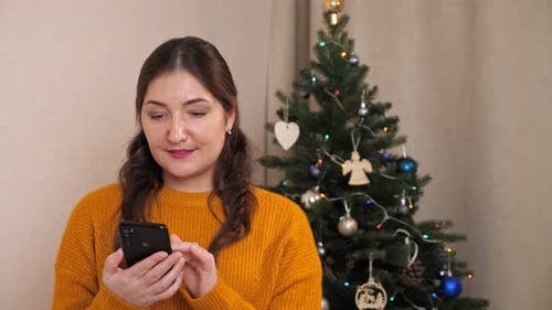 Woman Using Smartphone by Christmas Tree Indoors