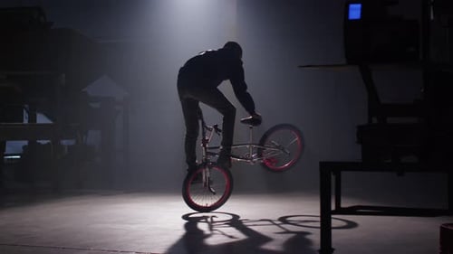 BMX rider doing tricks in dark warehouse.