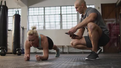 Fit caucasian woman performing plank exercise with male trainer holding digital tablet at the gym