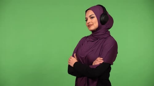 A Young Beautiful Muslim Woman Listens to Music with Headphones on and Smiles Green Screen