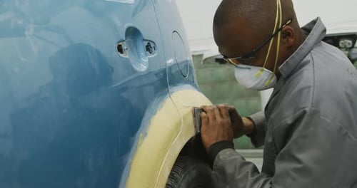 Afroamerikanischer Automechaniker, der eine Gesichtsmaske trägt und einen Polierer an der Seite eines Autos benutzt