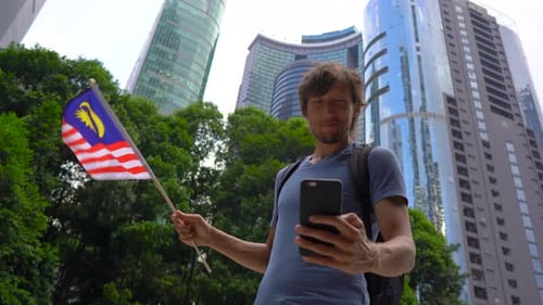Un jeune homme tient un smartphone et agite le drapeau malaisien avec des gratte-ciel en arrière-plan