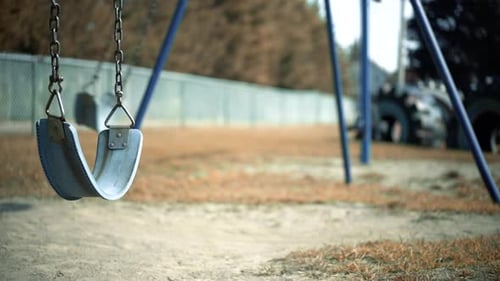 Verlassenes leeres Schaukelset Schaukeln im Wind auf Kinderspielplatz mit Zaunlinie im Hintergrund