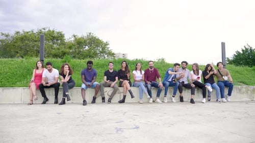Group of friends sitting in a row on wall in park with smartphones