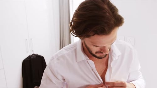 Man Buttons White Dress Shirt in Modern Bedroom