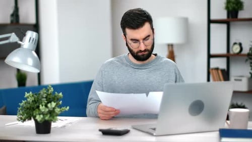 Man Reviews Documents at Desk with Laptop
