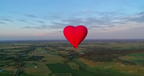 Flight of a red airship. Beautiful hot air balloon in heart shape travelling over the green fields i