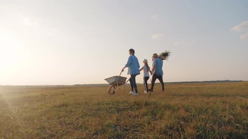 Young Family Having Fun Outdoors in Their Farm. Gardener Woman Pushing Wheelbarrow with Vegetables