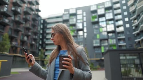 Formally Dressed Woman Standing in a Business District with Coffee in Hand and Using a Smartphone