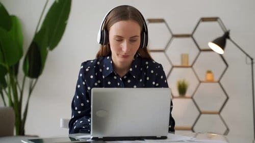 A Young Female Project Manager Puts on Headphones and Works on a Laptop Computer in an Office