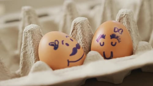 Video of close up of two brown eggs with drawn faces in egg carton background