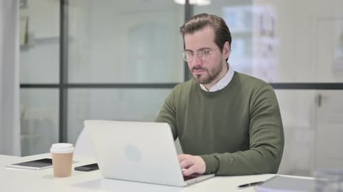 Junger Geschäftsmann lächelt in die Kamera, während er Laptop im Büro benutzt
