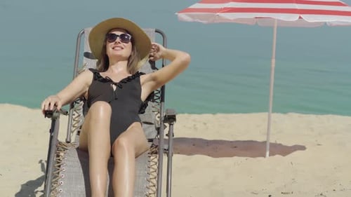 Happy Beautiful Young Woman in Swimsuit and Sunglasses Resting on Sun Lounger on Sandy Beach