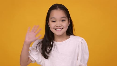 Cute Friendly Asian Girl Waving Hand Hi and Smiling To Camera Orange Studio Background
