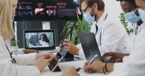 Médicos con máscaras sentados en el lugar de trabajo conjunto y viendo una videoconferencia