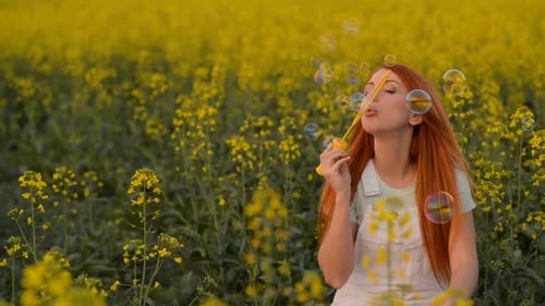 Young Redhair Woman Blowing Bubbles at the Camera Outdoors in Summer Meadow