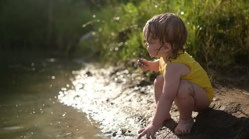 Little Funny Cute Blonde Girl Child Toddler in Yellow Wet Bodysuit Playing By the Lake Waterside