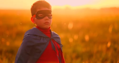 A Child in the Costume of a Superhero in a Red Cloak Runs Across the Green Lawn Against the Backdrop