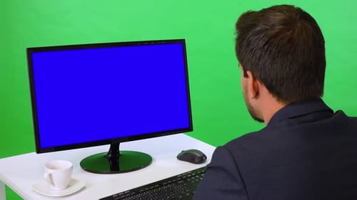 A Young Businessman Sits at a Desk and Looks at a Desktop Computer - Closeup - Green Screen Studio