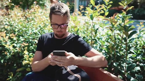 Man Is Browsing Social Nets By His Smartphone Outdoors in Park, Sit on Bench