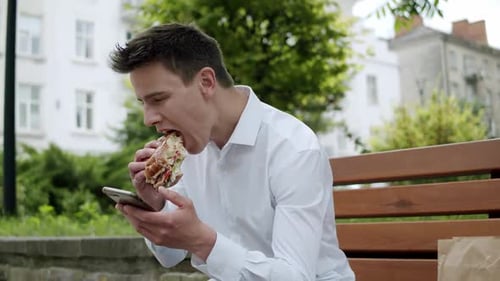 Young Man Typing on Smartphone and Eating a Burger When Sitting on Street Bench