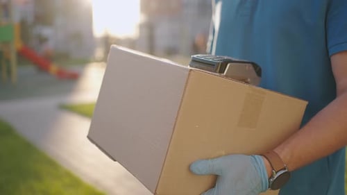 A Male Deliveryman in Gloves Brings a Box and Gives the Terminal to Pay for the Purchase