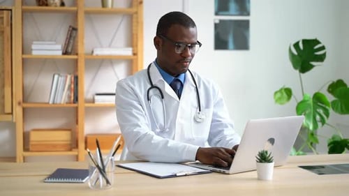 African American Male Doctor is Working with Laptop and Writing at Table in Clinic Spbas